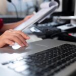 A close-up of a hand using a laptop touchpad with papers and a keyboard in the background.